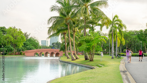 Beautiful garden in Chatuchak park Bankok Thailand, fresh green grass lawn yard under coconut palm trees beside a lake, orange concrete arch curve bridge, people running in good care landscapes