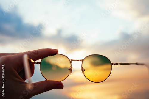 light skinned hand holds metal round sunglasses through which you can see a beautiful sea sunset in the mountains of blue and orange colors