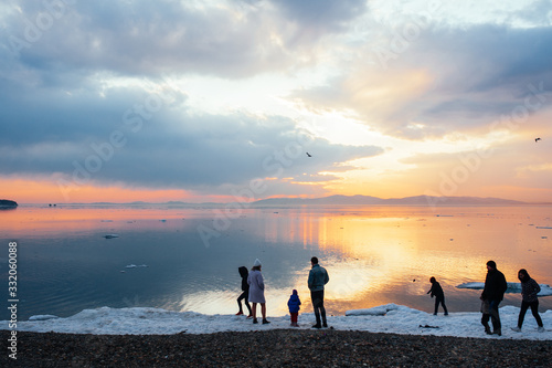 Silhouettes of people walking by the sea with the edge of the ice and admiring the incredibly beautiful orange and blue sunset with clouds and rays of the sun.