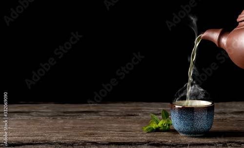 Hot green tea is poured from the teapot into the blue bowl, vintage wooden table, steam rises above the cup. Tea leaves next to the cup. Close-up, tea ceremony, minimalism, copy space for text.