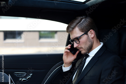 Handsome businessman talking with phone sitting with laptop on the backseat of the car.