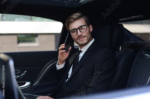 Handsome businessman talking with phone sitting with laptop on the backseat of the car. © ty