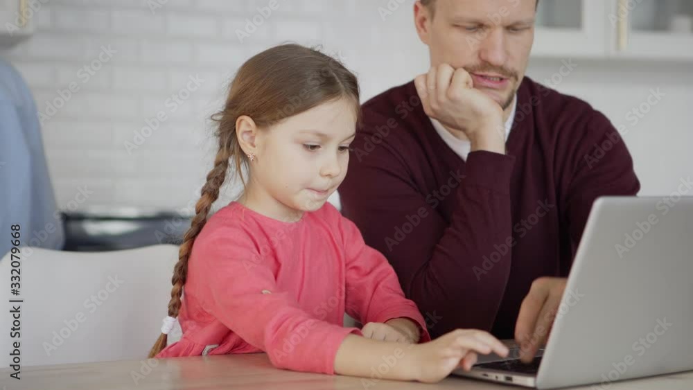 Tilt down medium shot of middle aged father teaching curious little daughter how to use laptop computer sitting at table at home Tilt down medium shot of middle aged father teaching curious little daughter how to use laptop computer sitting at table at home
