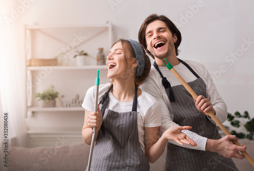 Murais de parede Cheerful Couple Using Mop Handles As Microphones, Having Fun During Spring-Clean