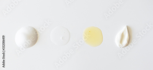 Sample beauty products for facial skin care on white background, above. Cleanser foam, tonic, oil serum essence drop and moisturizer cream smear