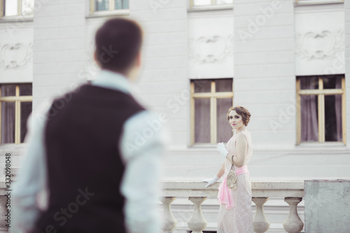 A beautiful woman in a retro dress stands against the background of a light building . She looks at the man and waits for him to approach her. Historical reconstruction photo