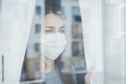 Young Woman in Face Mask Looking out the Window. Staying Home in Quarantine.