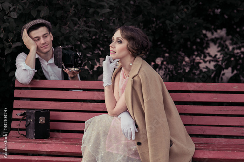 A young couple is photographed on a retro camera . A woman sits thinking on a bench , a man thoughtfully photographs a stylishly dressed woman in a retro style.Historical reconstruction photo