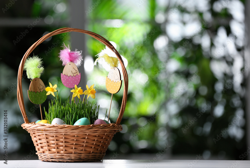 Wicker basket with colorful Easter eggs, wooden figures and flowers on white wooden table against blurred green background. Space for text Wicker basket with colorful Easter eggs, wooden figures and flowers on white wooden table against blurred green background. Space for text