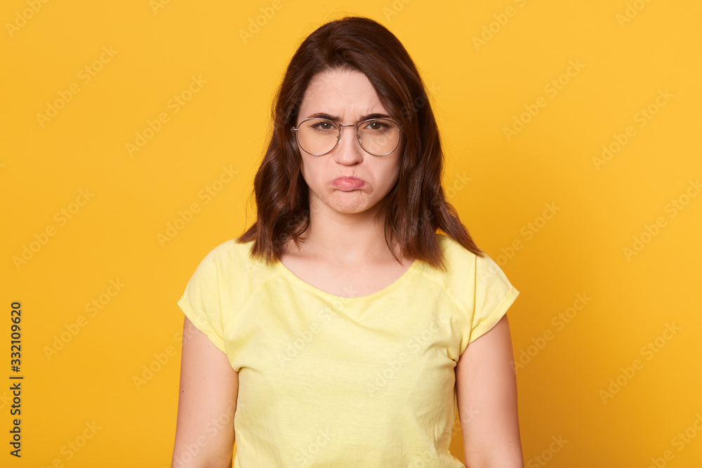 Close up portrait of beautiful young offended lady weraring casual t shirt and glasses, posing isolated over yellow background, woman expressers sadness, looks unhappy. People emotions cocnept. Close up portrait of beautiful young offended lady weraring casual t shirt and glasses, posing isolated over yellow background, woman expressers sadness, looks unhappy. People emotions cocnept.