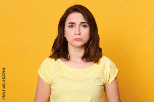 Picture of upset sad miserable young woman having frowned facial expression, standing isolated over yellow background in studio, being unsatisfied, having bad luck. People and bad mood concept.