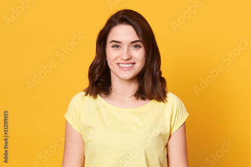 Canvas Print Image of cheerful positive good looking young female smiling sincerely, having pleasant facial expression, looking directly at camera, wearing yellow t shirt, standing isolated over yellow background