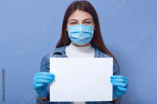 Panic woman with blankpaper for some inscription standing isolated over blue background, young female wearing demin jacket, flu disponsable medical mask and protective gloves. Coronavis, covid concept photo