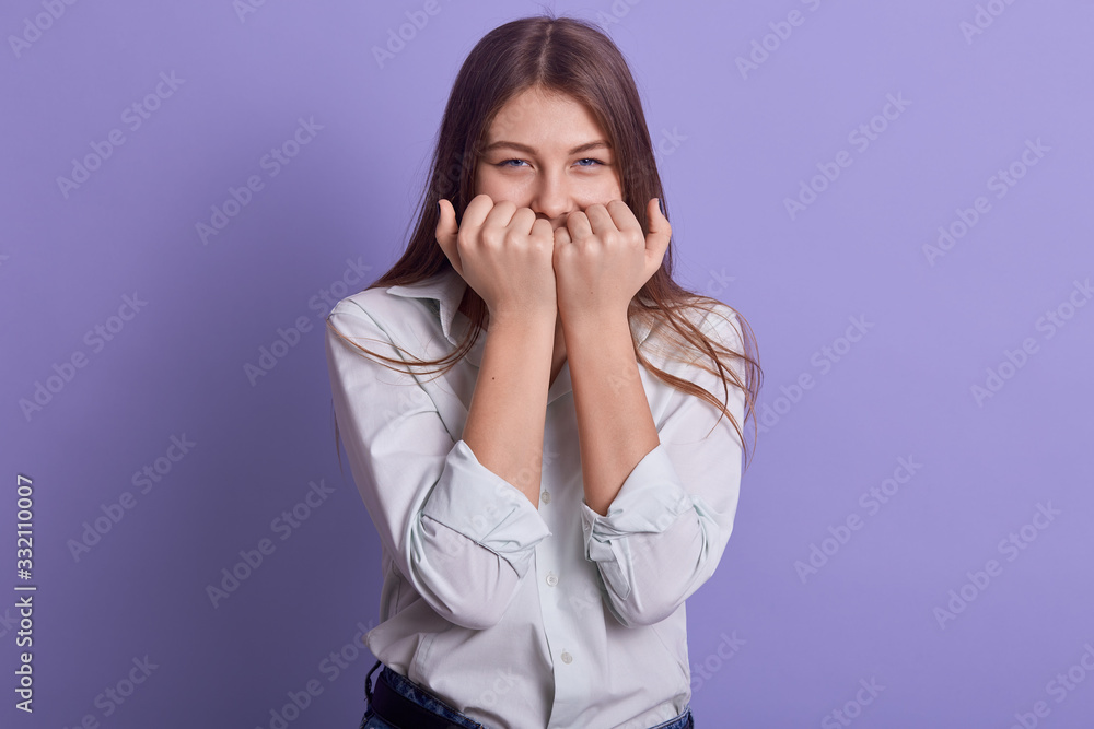 Horizontal indoor shot of attractive beautiful young female covering her mouth with both fists, standing isolated over lilac background in studio, having cunning facial expression. Youth concept. Horizontal indoor shot of attractive beautiful young female covering her mouth with both fists, standing isolated over lilac background in studio, having cunning facial expression. Youth concept.