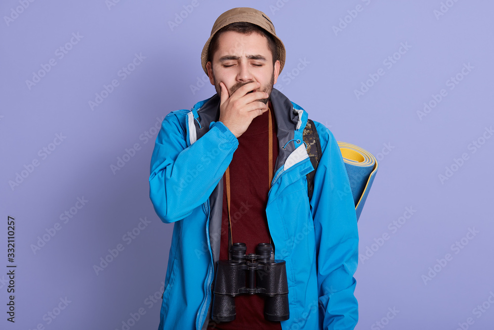 Horizontal shot of young explorer closing his eyes, covering his mouth with hand and yawning, stands with binoculars, having sleeping pad aback, wearing casual outfit. Travelling, lifestyle concept. Horizontal shot of young explorer closing his eyes, covering his mouth with hand and yawning, stands with binoculars, having sleeping pad aback, wearing casual outfit. Travelling, lifestyle concept.
