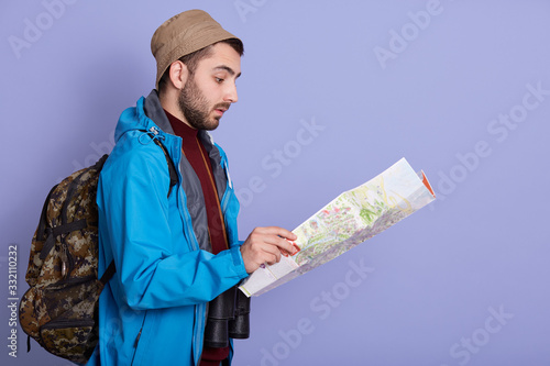 Profile of attentive serious backpacker looking for trip way, being alone, holding map, choosing route, wearing backpack, hat and jacket, having binoculars around neck. Copyspace for advertisement. photo