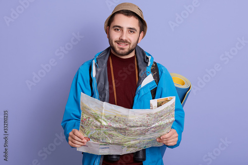 Portrait of handsome charismatic young tourist holding map in both hands, spending his free time in active way, wearing sweatshirt, jacket and hat, having sleeping pad aback. Orientation concept. photo