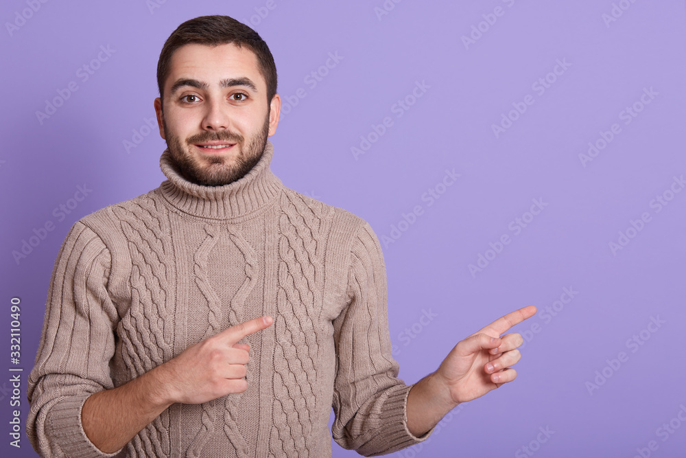 Cloe up portrait of handsome man dresses beige sweater looks curiously directly at camera, pointing with index fingers aside, showing free space for your advertising content, isolated over lilac wall. Cloe up portrait of handsome man dresses beige sweater looks curiously directly at camera, pointing with index fingers aside, showing free space for your advertising content, isolated over lilac wall.