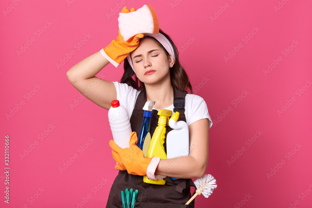 Indoor shot of young tired housewife wearing brown apron, white casual t shirt, haedband, posing with cleaning deretgents isolated over pink background, maid touching her forehead, holds sponge. Indoor shot of young tired housewife wearing brown apron, white casual t shirt, haedband, posing with cleaning deretgents isolated over pink background, maid touching her forehead, holds sponge.