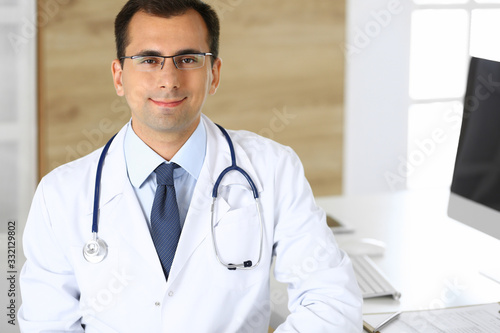 Doctor man sitting at the desk at his working place and smiling at camera. Perfect medical service in clinic. Happy future of medicine and healthcare