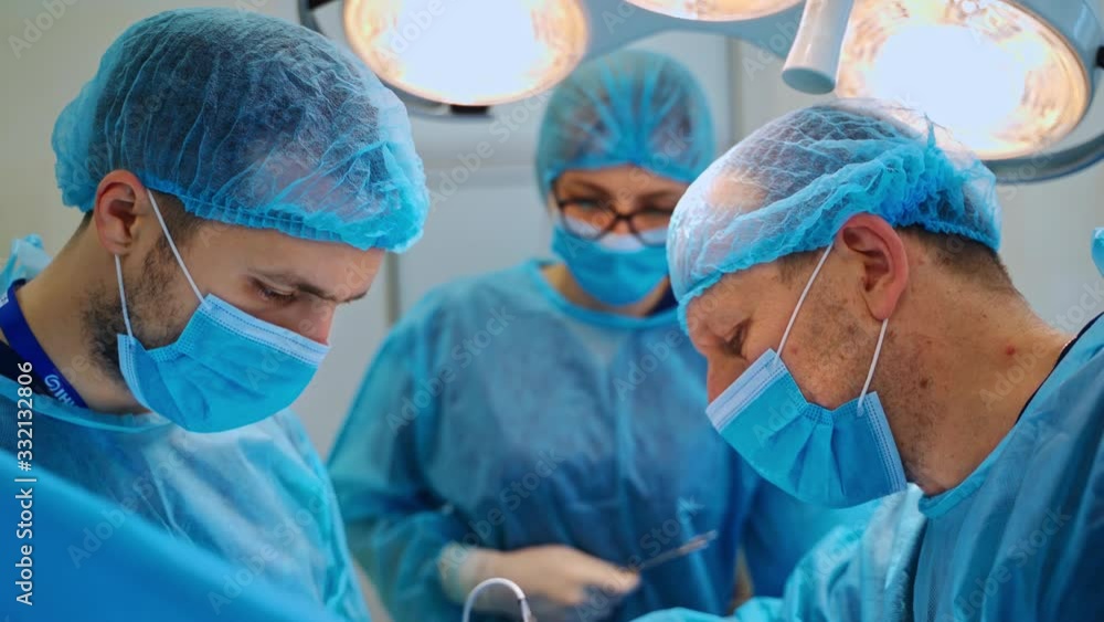 Two male specialists and assistant perform a surgery. Professional surgeons in blue uniform use medical instruments during an operation. Two male specialists and assistant perform a surgery. Professional surgeons in blue uniform use medical instruments during an operation.