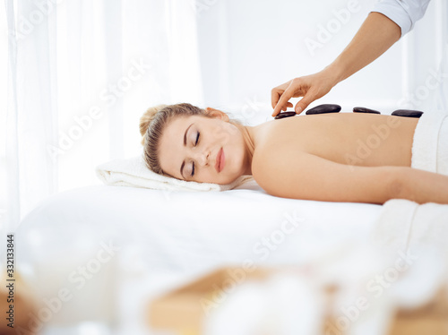 Young and blonde woman enjoying treatment with hot stones in spa salon. Beauty concept