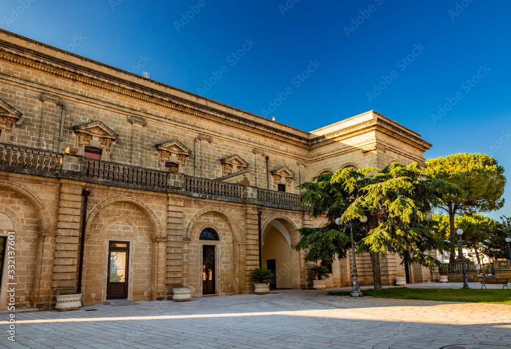 Acquarica del Capo, Presicce, Salento, Puglia, Italy. Town square with the seat of the municipality and the town hall. The ancient stone and brick building. Flower beds with the lawn and palm trees.