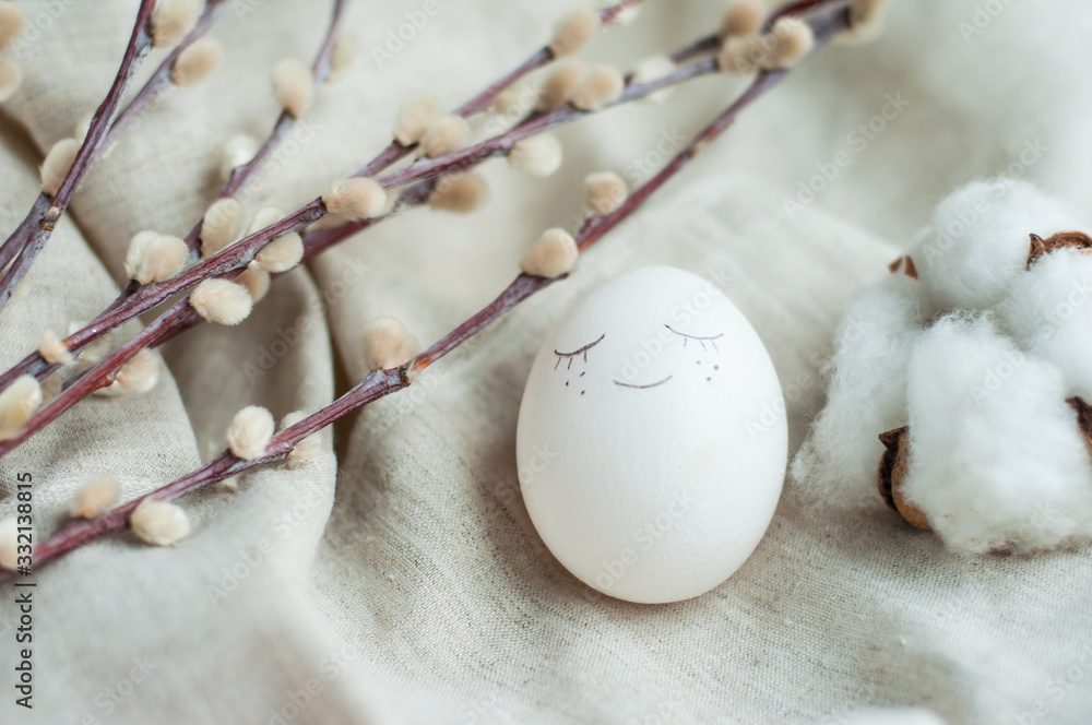 white Easter egg with a face. Next to it is a willow branch and flax on a gray linen background