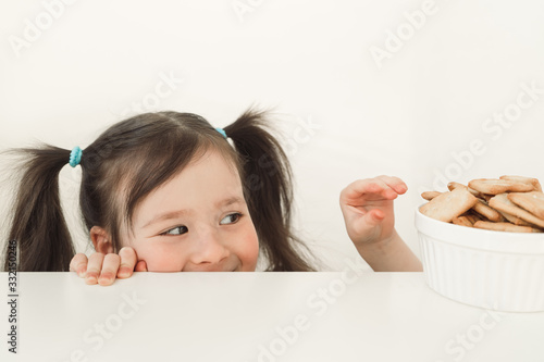 A girl peeks out from the table. The child wants to steal cookies. Funny baby hunts for delicious cookies