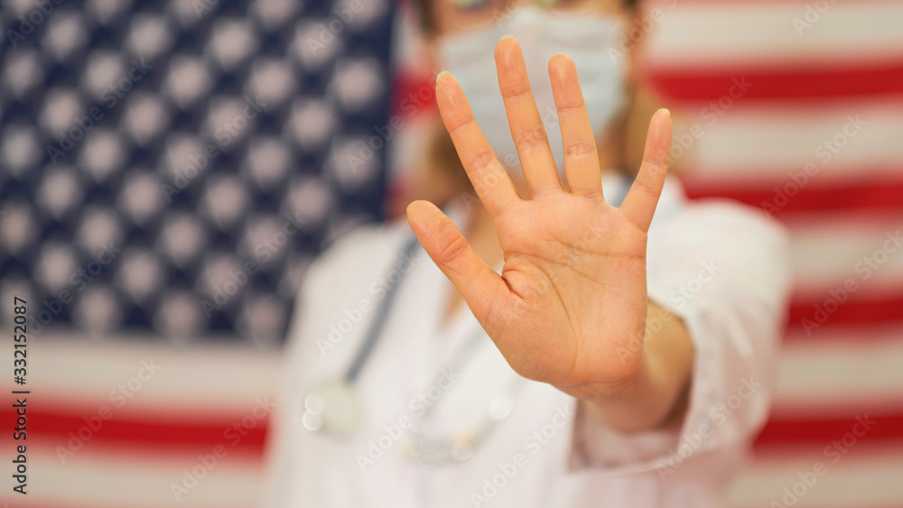 Doctor stands against the backdrop of the USA flag and shows a stop sign with his hand. Novel coronavirus 2019-nCoV with text on blue background. Virus Pandemic Protection Concept