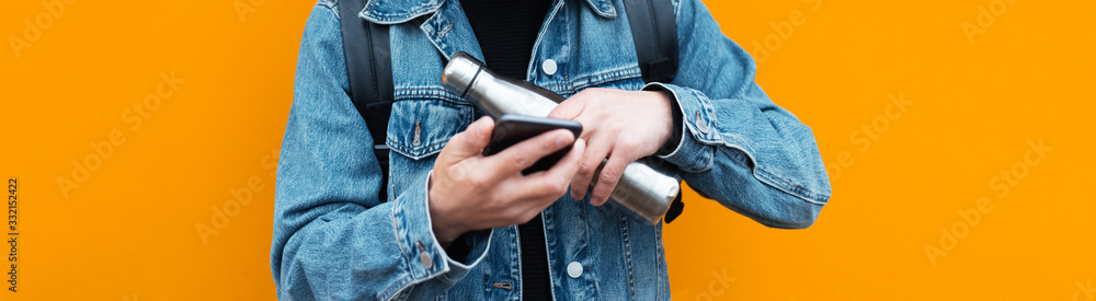 Close-up of guy in denim jacket holding reusable eco, steel stainless thermo water bottle, using smartphone, on background of orange color. Panoramic banner concept. Close-up of guy in denim jacket holding reusable eco, steel stainless thermo water bottle, using smartphone, on background of orange color. Panoramic banner concept.