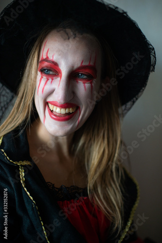 Portrait of a mysterious girl with fair hair as a witch with bright make-up and leaking lipstick mysteriously looks to the side with wide smile