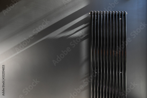 Stylish solution of the house heating system design. Vertical long heater radiator mounted on a black wall, casts deep shadows on a Sunny day