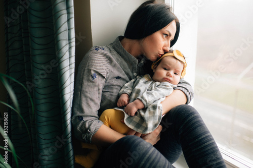 Pretty woman with black hair sits on a windowsill with her pretty little child and smiles