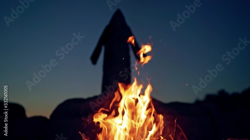 Scary silhouette od black figure on the hill behind fire. Orange flame at night and horrorful death in black cloak with hood is waving arms. Halloween concept. photo