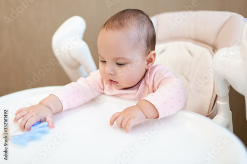 Little toddler is playing at the table. A girl sits in a highchair and reaches for a rattle. Baby is playing.
