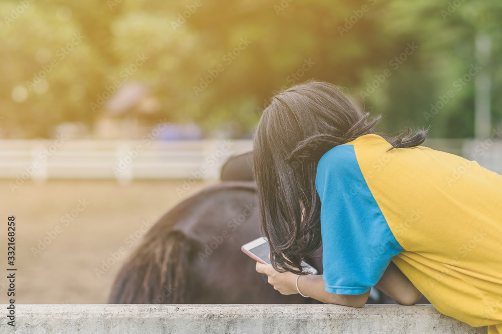 Girl in yellow and blue shirt use smartphone to take photos at the horse riding field in the evening.