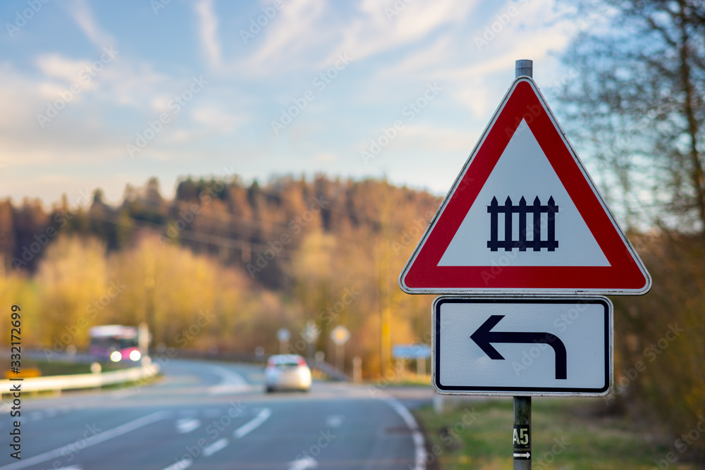 A red and white triangular traffic sign warns of a railway crossing if you turn left. In the background a street with a car and bus. It is a late winter afternoon in Germany. A red and white triangular traffic sign warns of a railway crossing if you turn left. In the background a street with a car and bus. It is a late winter afternoon in Germany.