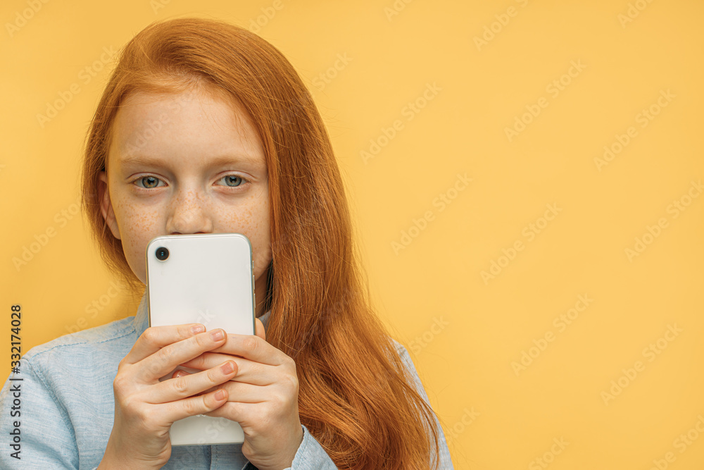 close-up portrait of little beautiful caucasian girl with white smartphone isolated over yellow background. red haired little child look at camera through mobile phone close-up portrait of little beautiful caucasian girl with white smartphone isolated over yellow background. red haired little child look at camera through mobile phone