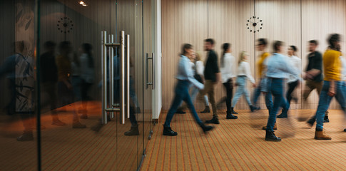 Group of office employees in motion go around at coworking space. People in business center walking  at hall. Motion blur. Wide image