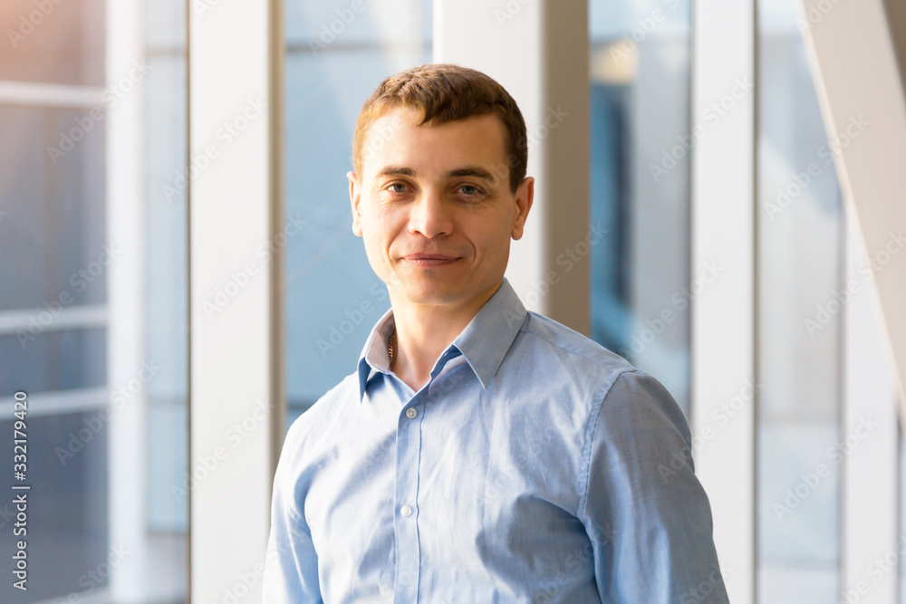 A 35-40 year old man in a blue shirt poses in office back hallway against the background of a large window. A 35-40 year old man in a blue shirt poses in office back hallway against the background of a large window.