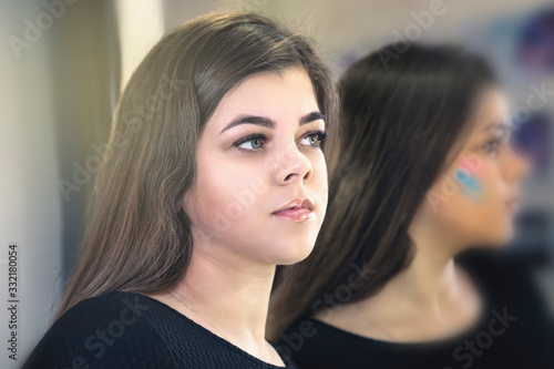 Portrait of a young beautiful girl near the mirror. Girl and her reflection in the mirror. The girl looks at the camera through the mirror. Selective focus.