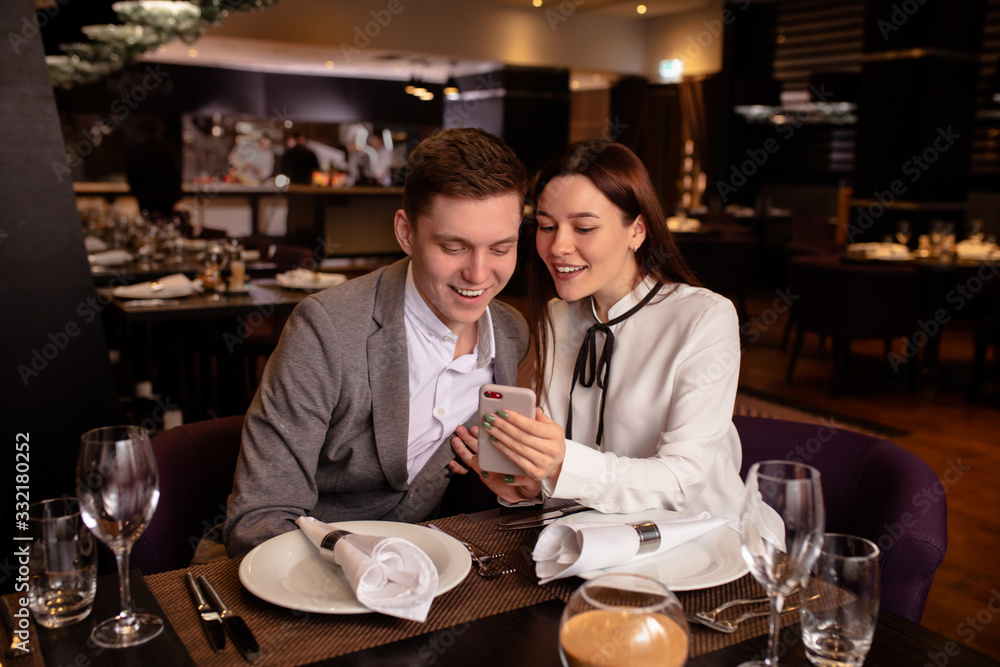 young caucasian couple have dinner together, look at smartphone in restaurant. woman show cool video to boyfriend. indoors. man and woman enjoy spending time together