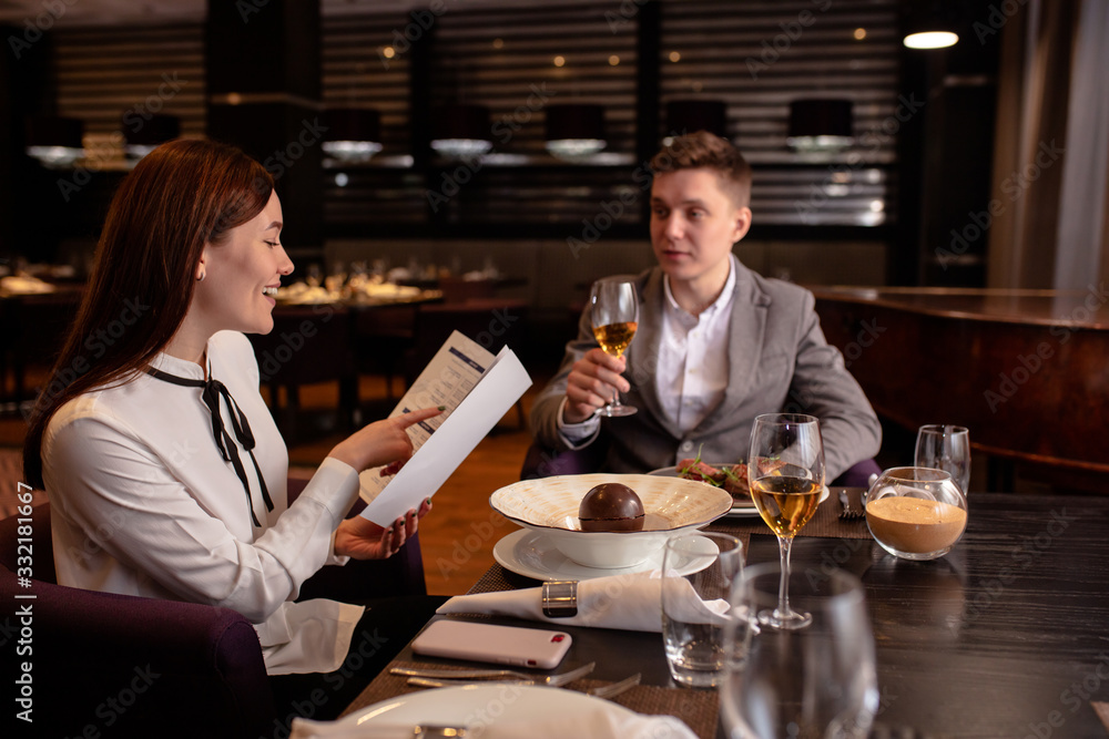 happy young caucasian couple spend their evening in restaurant, man adn woman on a date. lovely lady and handsome guy celebrate anniversary. woman looks at the menu, wants to order a delicacy