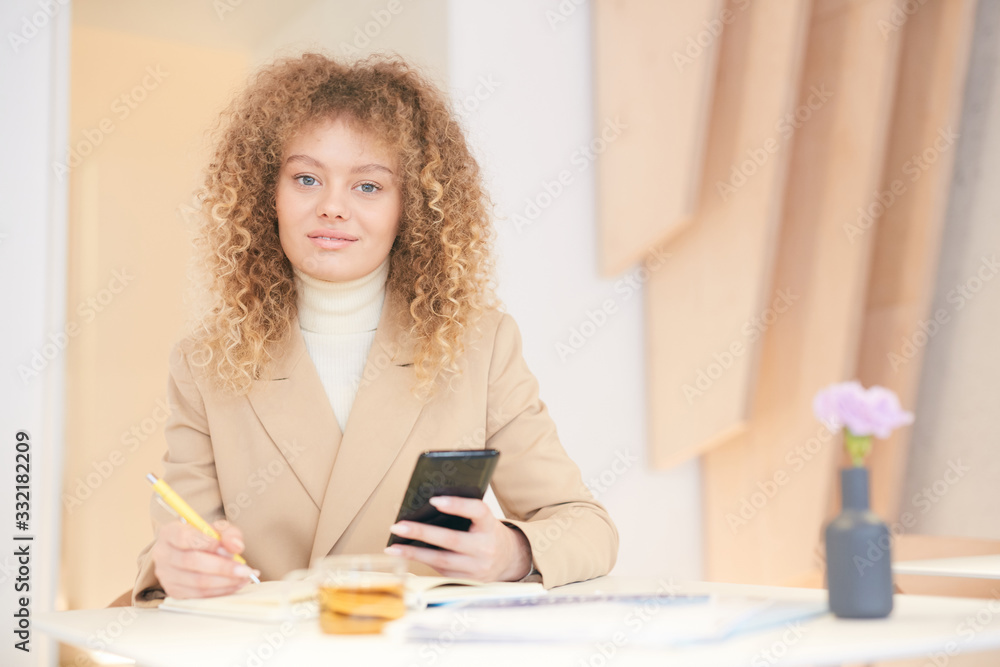 Warm toned portrait of curly haired young woman looking at camera and holding smartphone while working or studying sitting at table in cafe, copy space Warm toned portrait of curly haired young woman looking at camera and holding smartphone while working or studying sitting at table in cafe, copy space