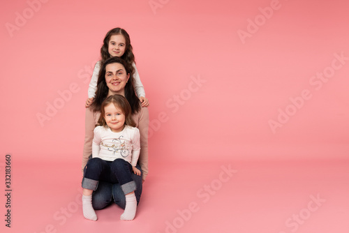 Beautiful young mother with her two daughters sitting on the ground one by one. Looking directly at the camera. Isolated on the pink background