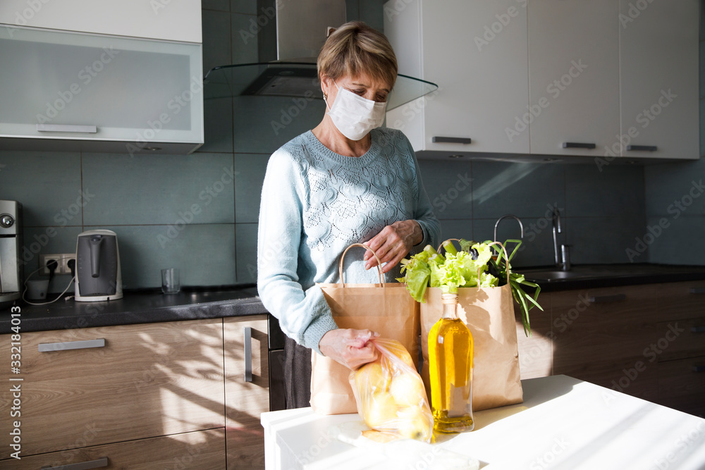 Seniors woman  wearing medical face mask with food bags at home in the kitchen. quarantine. health concept. Corona Virus. order of products online. Delivering products to home - obrazy, fototapety, plakaty 