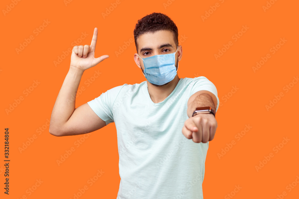 You are loser! Portrait of bossy angry man with medical mask in white t-shirt showing loser gesture and pointing at camera, mocking your defeat. indoor studio shot isolated on orange background