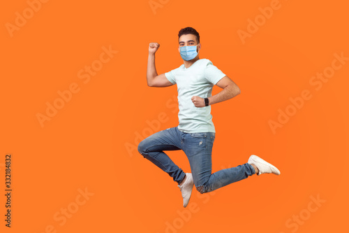 Full length of positive inspired brunette man with medical mask in sneakers  denim outfit jumping in air or running quickly fast. indoor studio shot isolated on orange background  empty copy space