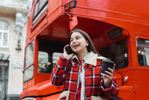 happy beautiful girl in checkered jacket talking on smartphone with coffee to go near red bus on city street